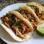 Three soft tortilla chicken tacos filled with shredded seasoned chicken and topped with fresh cilantro, served with a lime wedge.