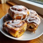 A stack of freshly baked cinnamon roll pastries generously topped with white icing, one is broken open.