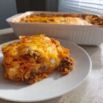 A generous serving of Dorito Casserole featuring ground meat, creamy sauce, and melted cheddar cheese, with the baking dish in the background.