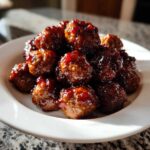 A close-up of a mound of glistening Grape jelly meatballs coated in a thick, dark, sweet glaze, served on a white plate.