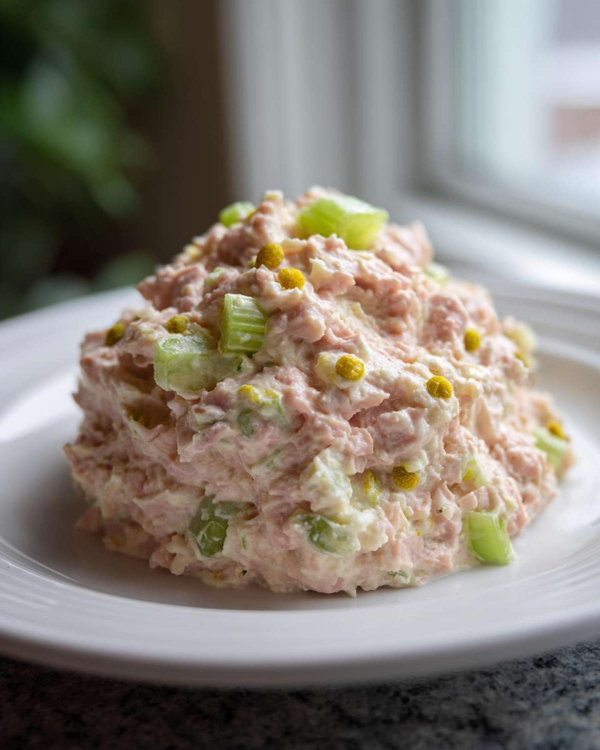 A mound of creamy ham salad recipe mixed with celery and yellow relish served on a white plate.