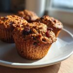 Close-up of four golden brown honey bran muffins topped with dried cranberries on a white plate.