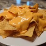 Close-up of tortilla chips generously covered with smooth, bright orange nacho cheese sauce on a white plate.