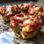 Close-up of one of the amazing pizza muffins with a bite taken out, showing the fluffy interior and melted cheese topping.