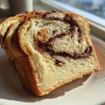 Two thick slices of homemade raisin bread featuring a visible cinnamon swirl, served on a white plate near a window.
