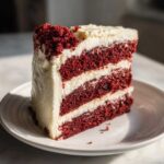 A tall slice of multi-layered red velvet cake with thick white cream cheese frosting, sitting on a white plate.