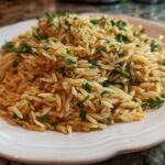 Close-up of a serving of fluffy, golden rice pilaf garnished with chopped fresh parsley on a white plate.