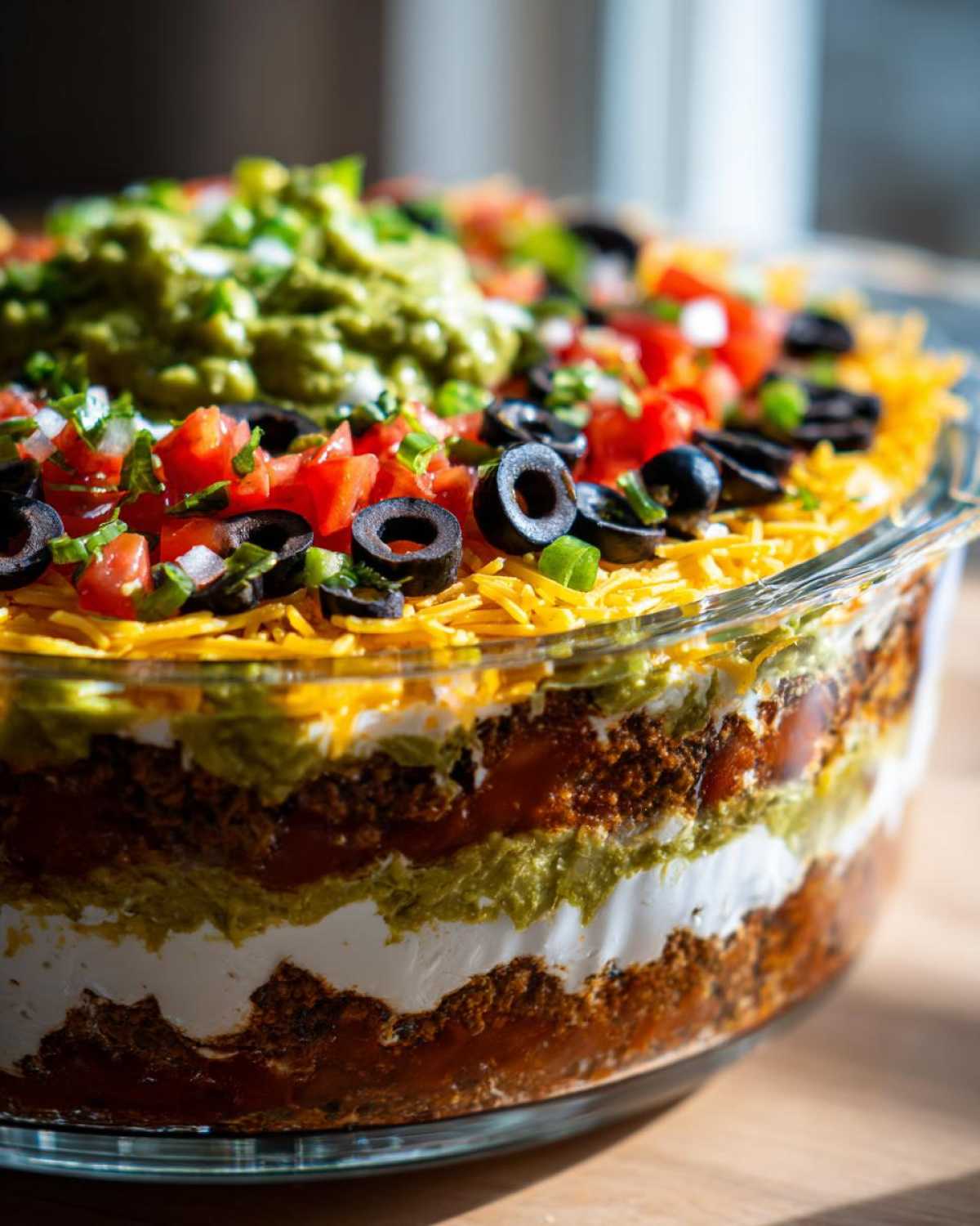 Close-up view of a colorful, layered Seven layer dip in a clear glass serving dish, topped with guacamole, tomatoes, and olives.