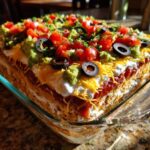 Close-up of a tall, layered Seven layer dip in a glass baking dish, topped with guacamole, tomatoes, olives, and cheese.