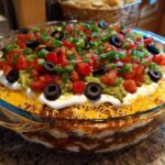 Close-up of a fully assembled Seven layer dip in a clear glass dish, topped with guacamole, sour cream, tomatoes, olives, and green onions.