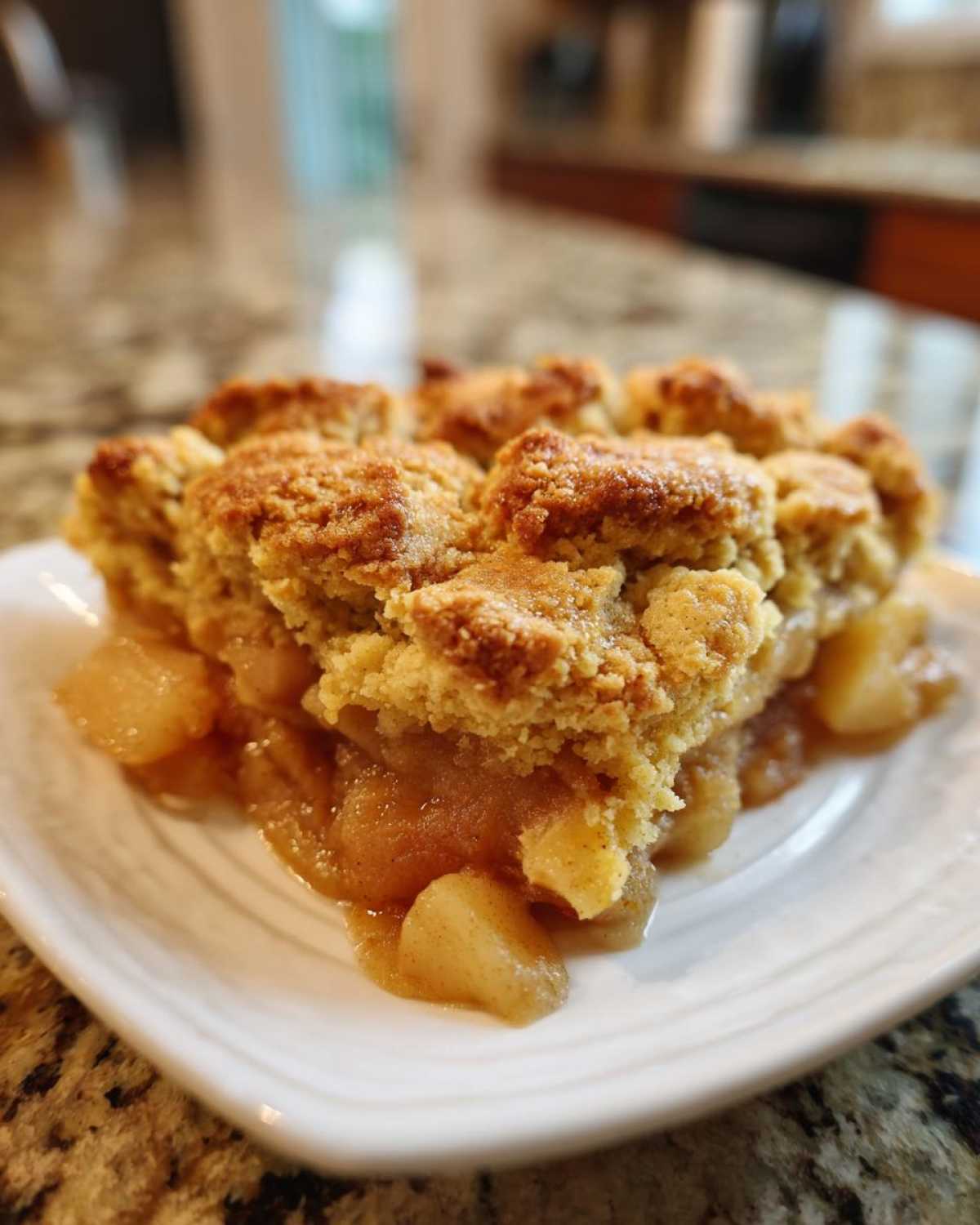 A close-up of a warm serving of apple cobbler recipe with a crumbly topping on a white plate.
