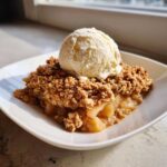 A warm slice of apple crisp recipe topped with a melting scoop of vanilla ice cream on a white plate.