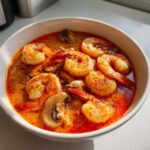 A close-up shot of a white bowl filled with vibrant red and orange tom yum soup, featuring several plump shrimp and sliced mushrooms.