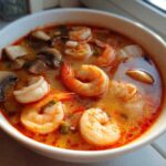 A close-up, vibrant bowl of authentic tom yum soup featuring large shrimp and sliced mushrooms in a rich, orange broth.