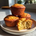 Four golden-brown banana protein muffins stacked on a plate, one is bitten open showing the moist interior.