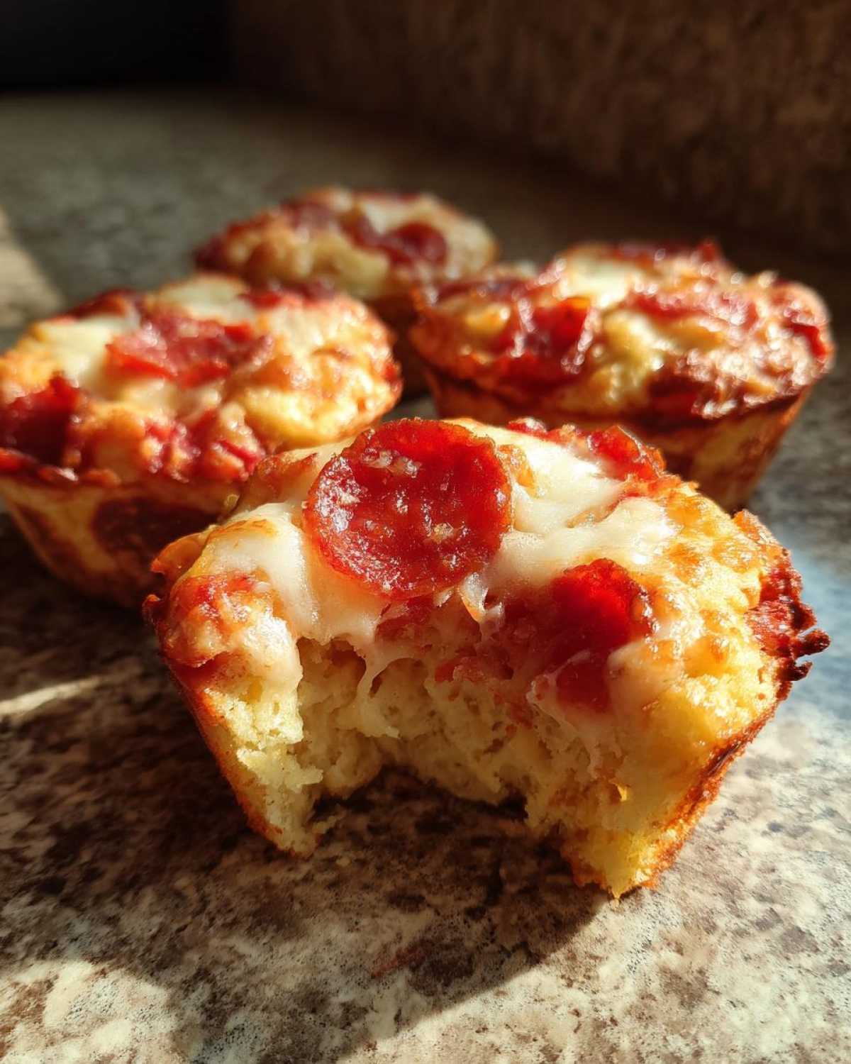 Close-up of a bitten pizza muffin topped with pepperoni and melted cheese, showing the fluffy interior of the pizza muffins.