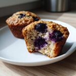 A close-up of a blueberry protein muffin cut in half showing the moist interior and burst blueberries, with another muffin behind it.