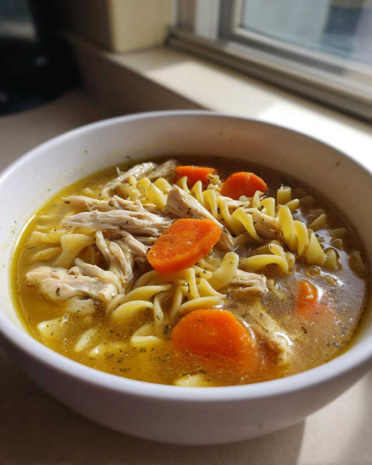 Close-up of a white bowl filled with rich chicken noodle soup, featuring shredded chicken, egg noodles, and bright carrot slices.