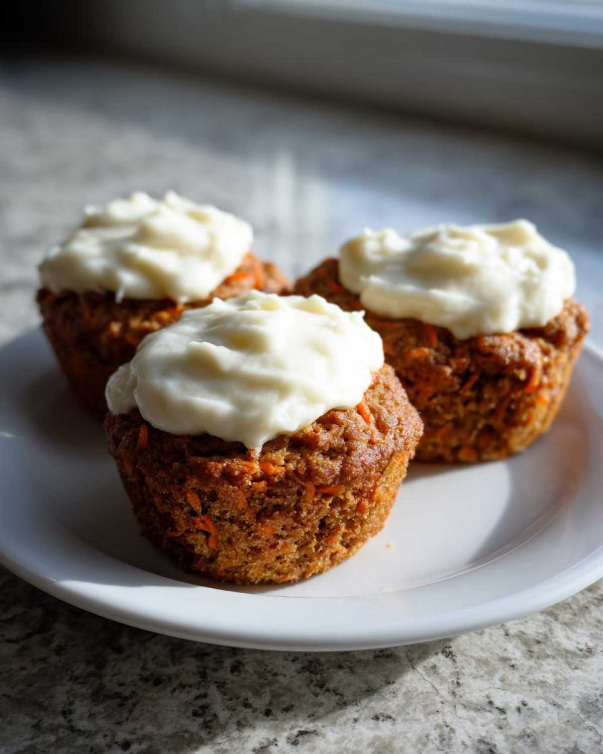 Three moist carrot cake muffins topped with thick cream cheese frosting, sitting on a white plate.