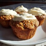 Four freshly baked carrot cake muffins topped with thick cream cheese frosting, sitting on a white plate.