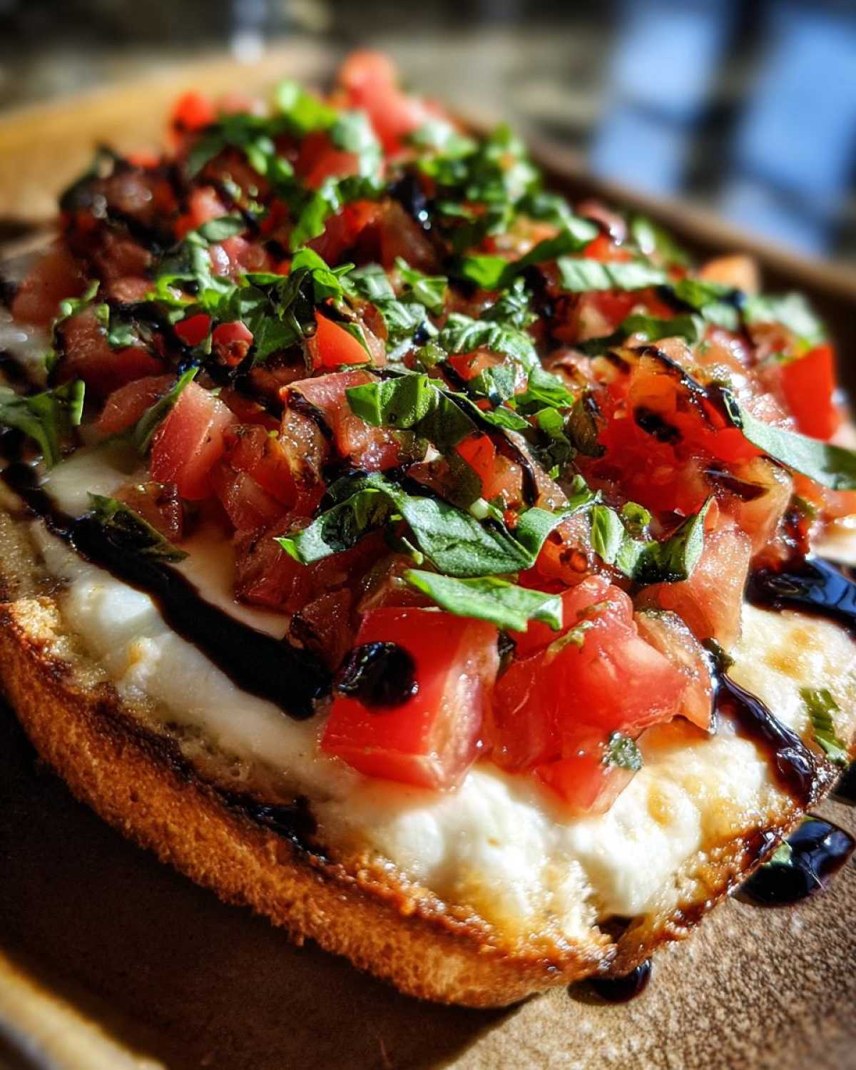 Close-up of toasted bread topped with melted cheese, diced tomatoes, fresh basil, and balsamic glaze, resembling a bruschetta dip serving.