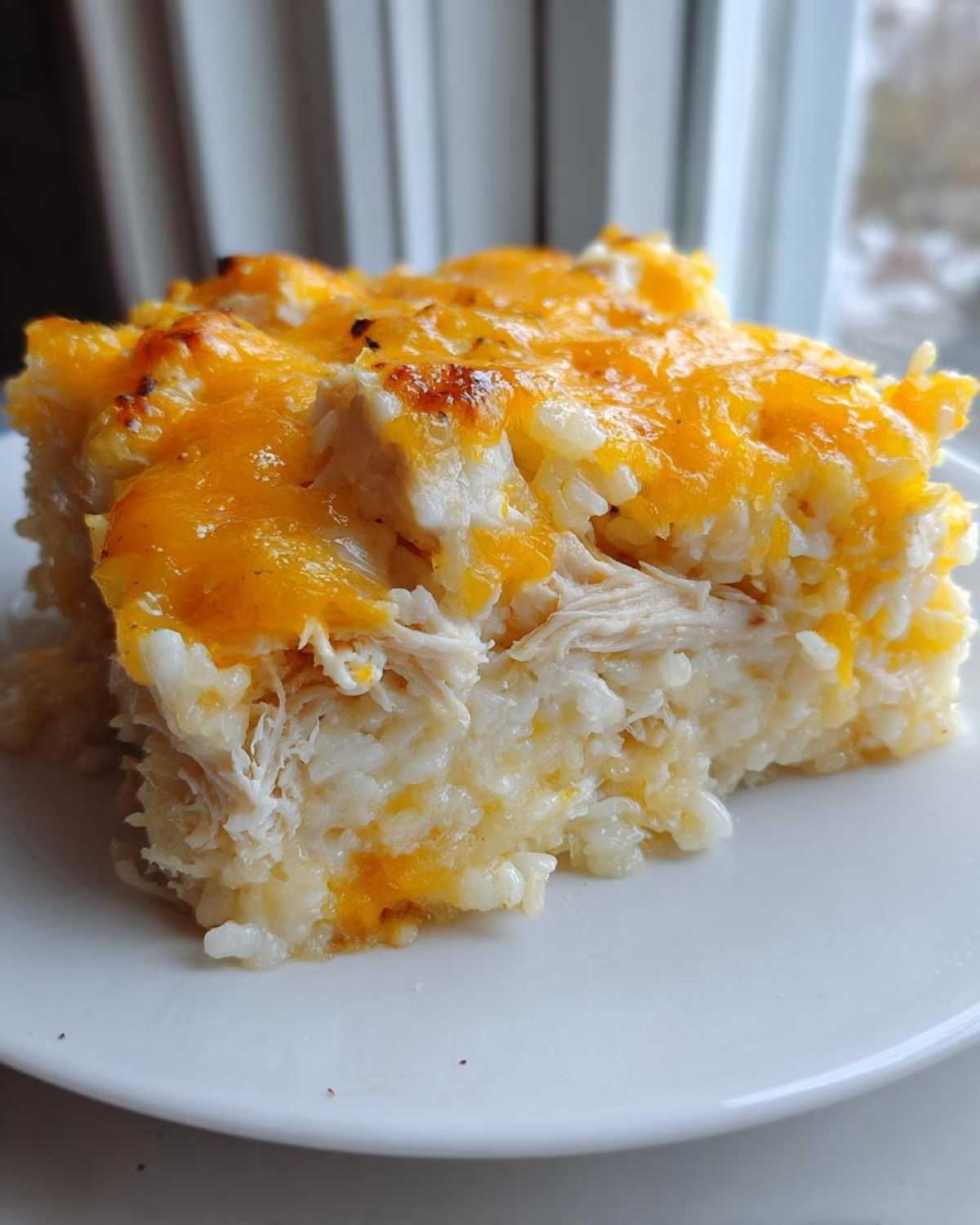 A square serving of cheesy chicken and rice casserole topped with melted cheddar cheese.