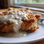 Close-up of crispy, golden brown chicken fried steak smothered in creamy, peppered white gravy on a white plate.