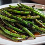 A close-up of bright green, blistered chinese green beans with garlic sauce piled on a white plate.