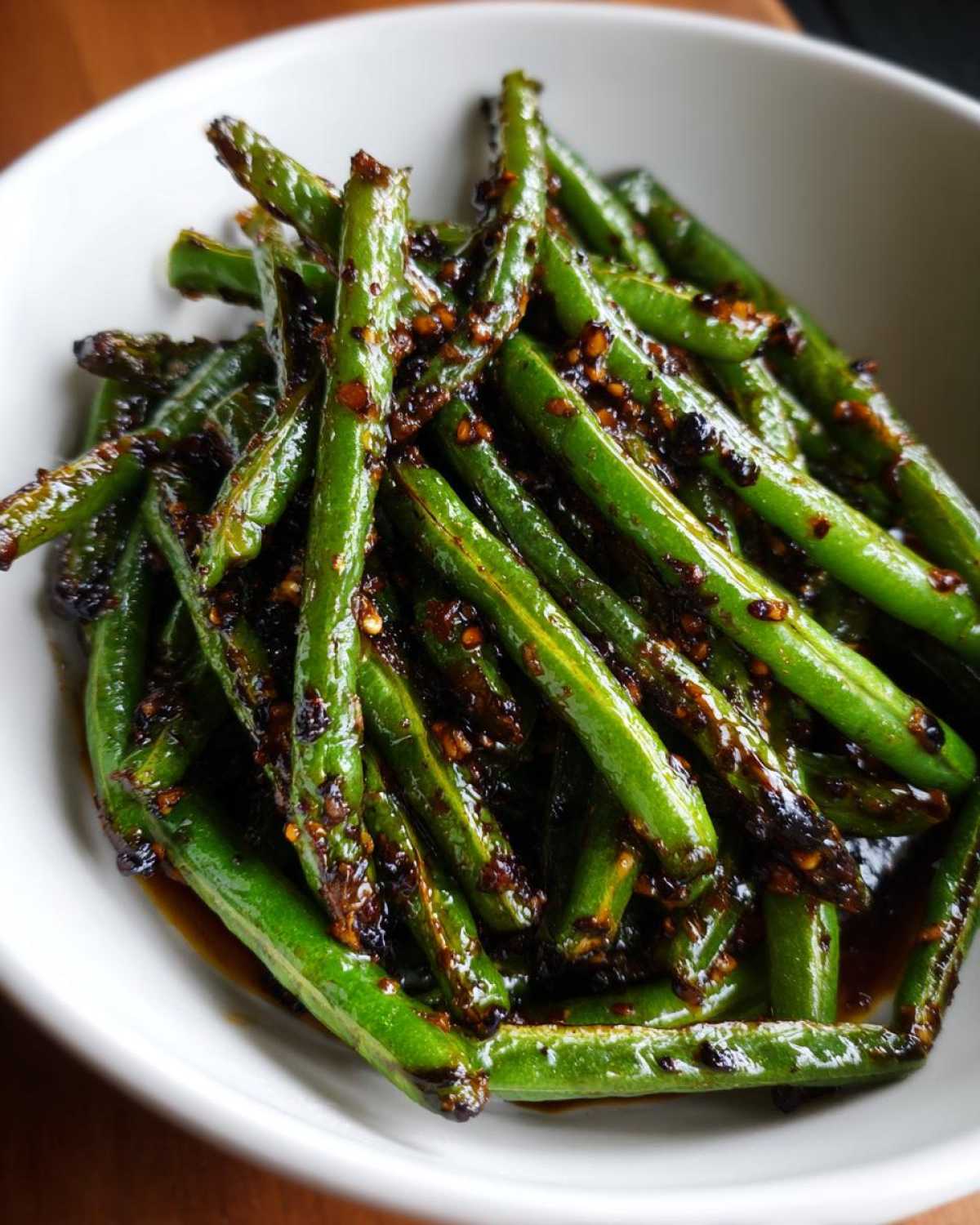Close-up of bright green beans coated in a dark, glossy chinese green beans with garlic sauce, served in a white bowl.