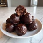 A stack of rich, chocolate-dipped Oreo balls on a white plate, one has a bite taken out showing the dark center.