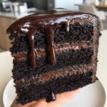 Close-up of a rich, dark slice of chocolate fudge cake layered with frosting and dripping with glossy chocolate ganache.