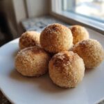 A close-up of five cinnamon sugar snowball cookies piled on a white plate, coated heavily in sugar.