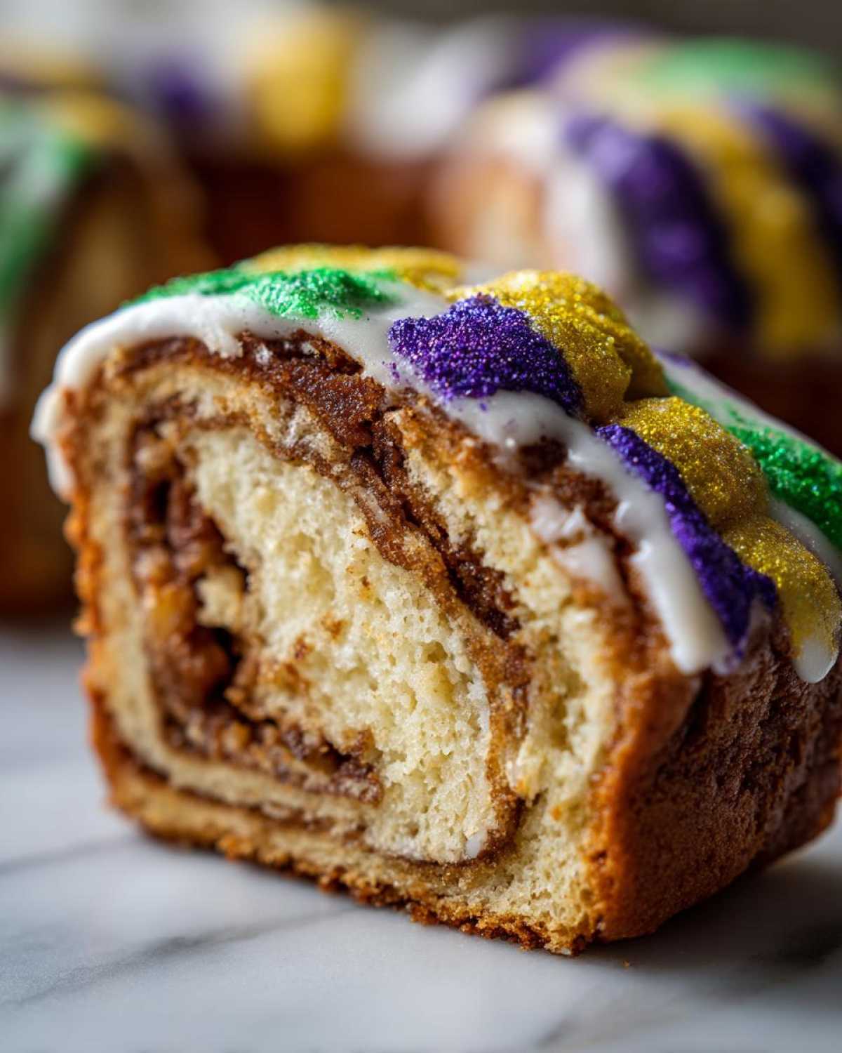 A close-up slice of a cinnamon swirl king cake topped with white icing and purple, gold, and green sanding sugar.