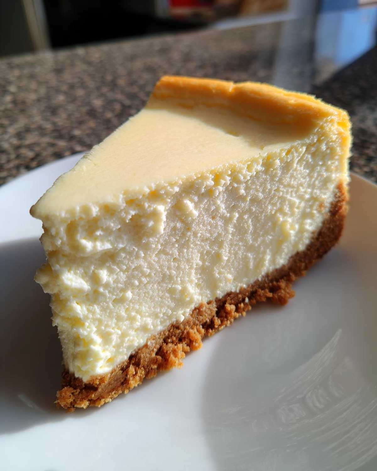 A close-up, sunlit slice of classic cheesecake recipe showing creamy filling and graham cracker crust on a white plate.