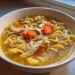 Close-up of a white bowl filled with homemade chicken noodle soup, featuring egg noodles, shredded chicken, and sliced carrots.