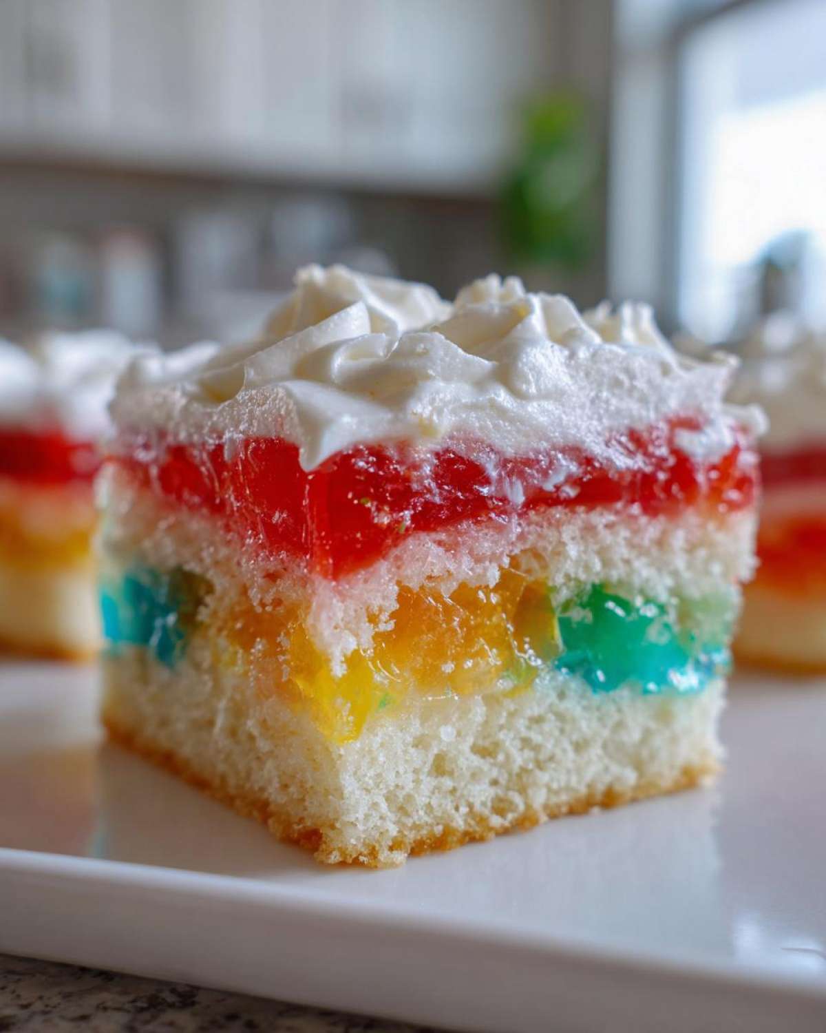 A close-up of a moist slice of 3-color jello poke cake showing colorful gelatin cubes soaked into the white cake layer, topped with whipped cream.