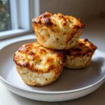 Three golden-brown cottage cheese egg cups starbucks hack stacked on a white plate near a window.