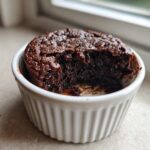 A close-up of a rich, dark chocolate Cottage Cheese Microwave Brownie served in a white fluted ramekin with a bite taken out.