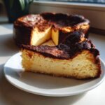 A close-up slice of creamy basque cheesecake with a dark, caramelized top sitting on a white plate, with the rest of the cake blurred in the background.