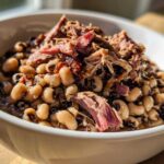 A close-up of creamy black eyed peas served in a white bowl, topped generously with shredded smoked pulled pork.