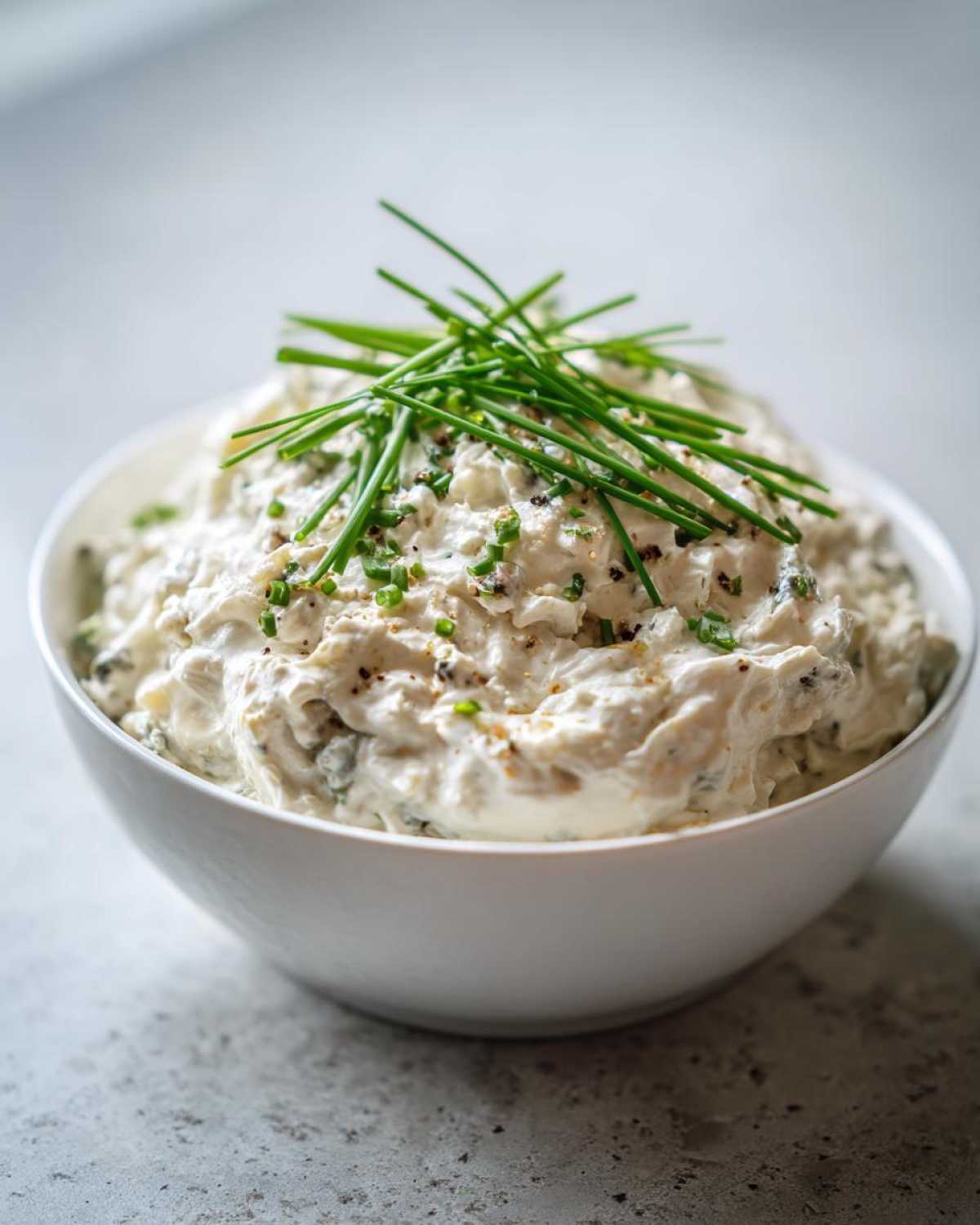 A close-up of creamy blue cheese dip in a white bowl, garnished heavily with fresh chives and cracked pepper.