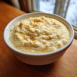 A white bowl filled with creamy, yellow cheese grits, lightly drizzled with butter and sprinkled with paprika.
