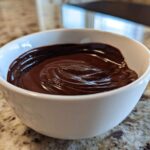 A close-up of rich, glossy chocolate dip swirled in a small white bowl on a granite countertop.