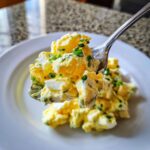 A spoonful of creamy, chunky egg salad recipe topped with fresh green chives, lifted over a white plate.