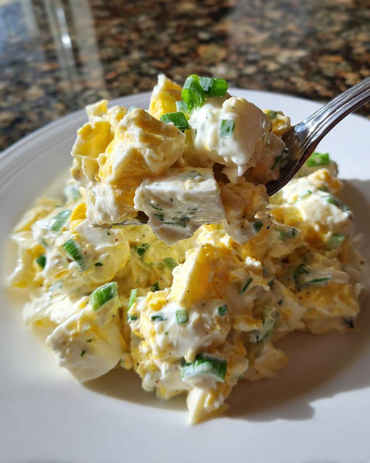 A spoonful of creamy egg salad recipe, featuring chopped eggs and green onions, lifted above a white plate.
