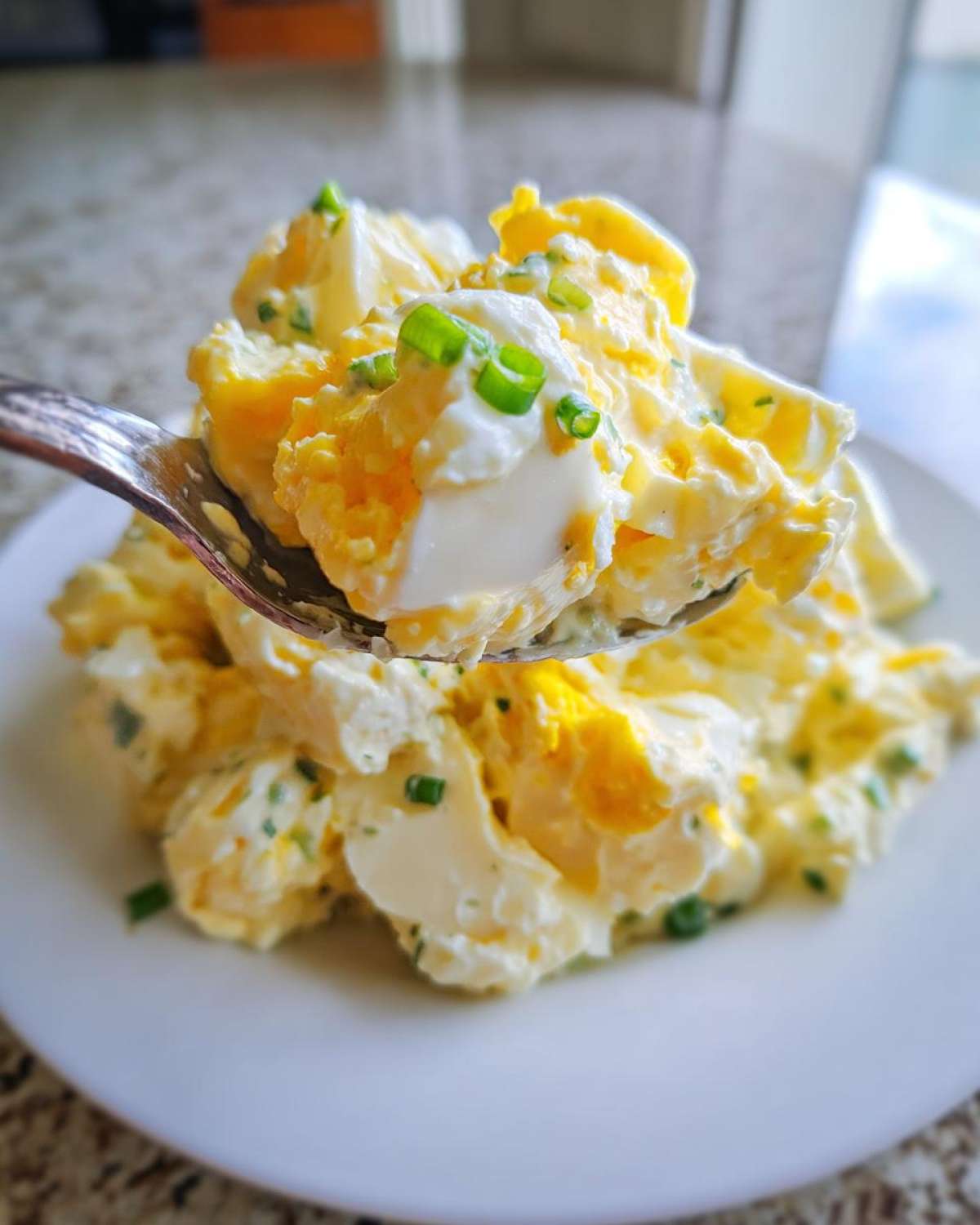 A spoonful of creamy egg salad recipe, topped with fresh green chives, lifted above a plate.