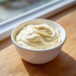 A white bowl filled with thick, creamy homemade mayonnaise sitting on a wooden surface near a window.