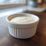 A small white ramekin filled with creamy, pale horseradish sauce sitting on a dark wooden table.