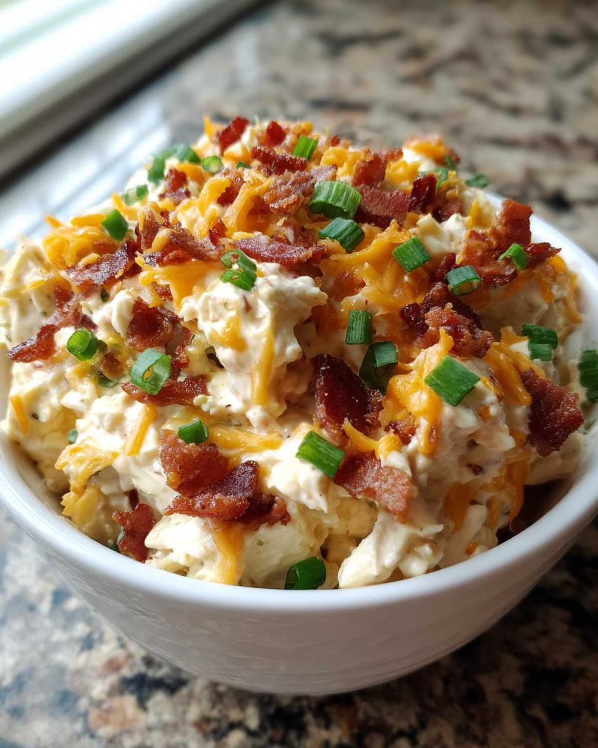 A close-up of creamy Million Dollar Dip served in a white bowl, topped generously with shredded cheddar cheese, bacon bits, and green onions.