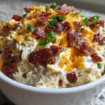 A close-up of creamy Million Dollar Dip topped generously with shredded cheddar cheese, crispy bacon bits, and green onions.